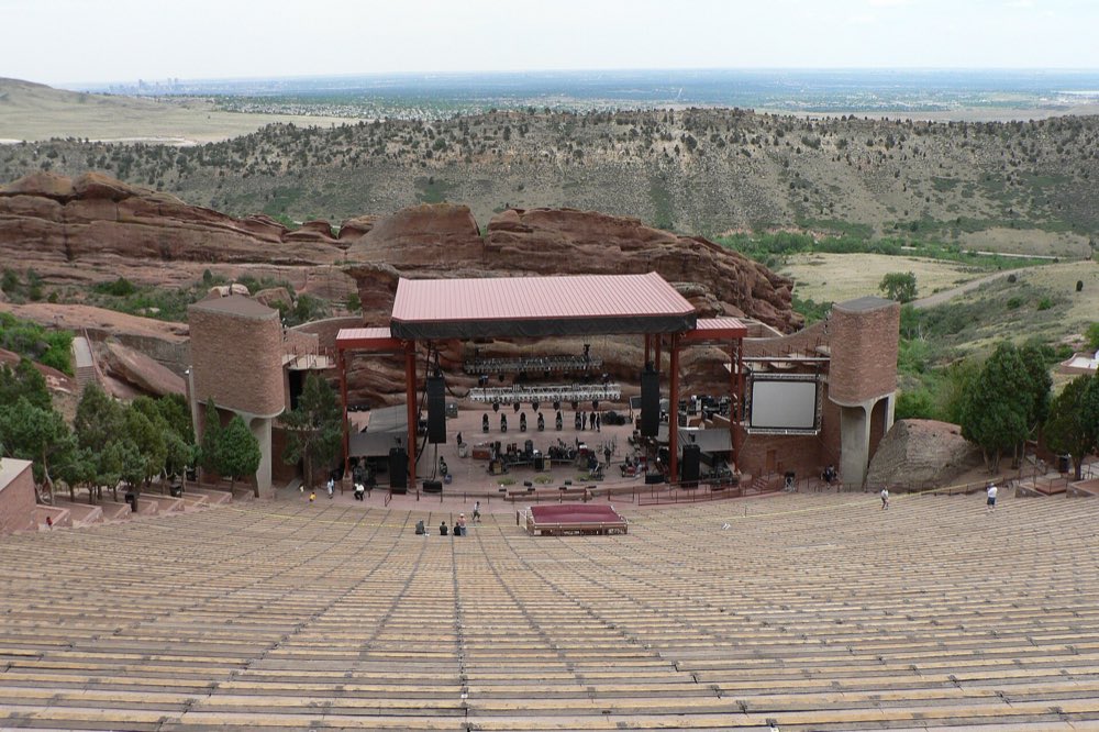 Rows of empty benches sloping down to the covered stage at Red Rocks Amphitheatre, framed by red sandstone, with the plains stretching to the horizon