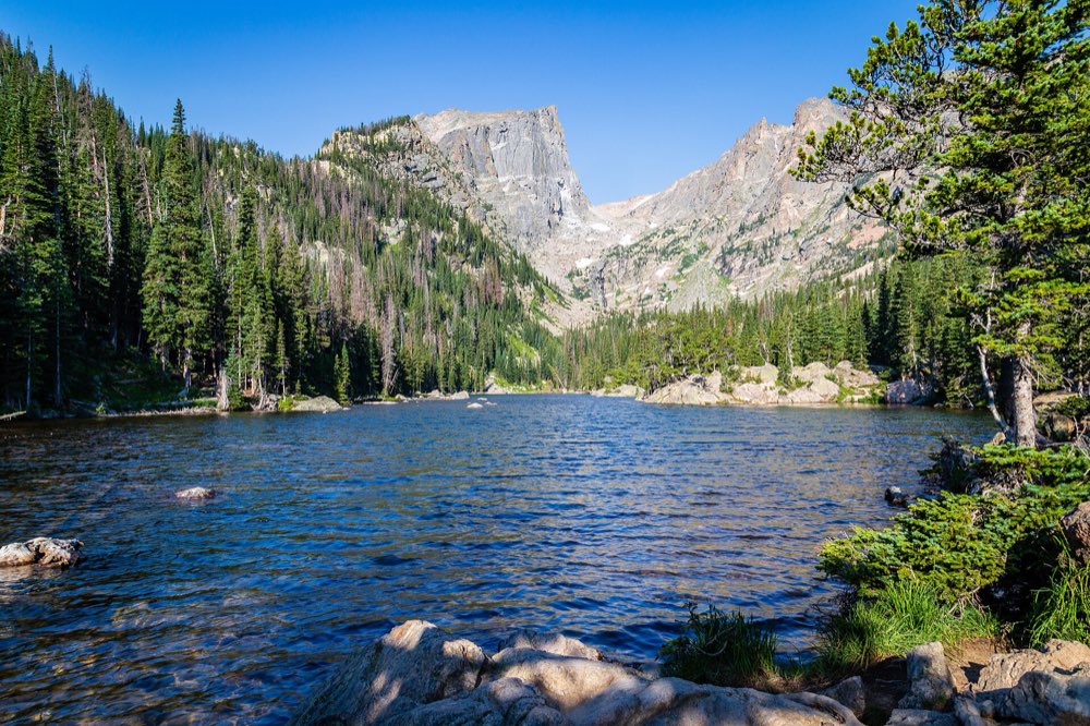 Dream Lake's clear blue water in morning sun, with the granite face of Hallett Peak rising above dark pines at the far end