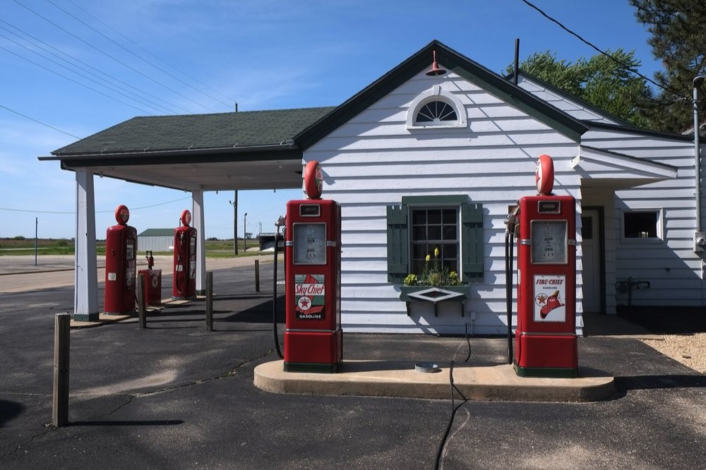 Two red vintage gas pumps in front of the white clapboard Ambler's Texaco station in Dwight, with a green roof, shuttered window and flower box under a deep blue sky