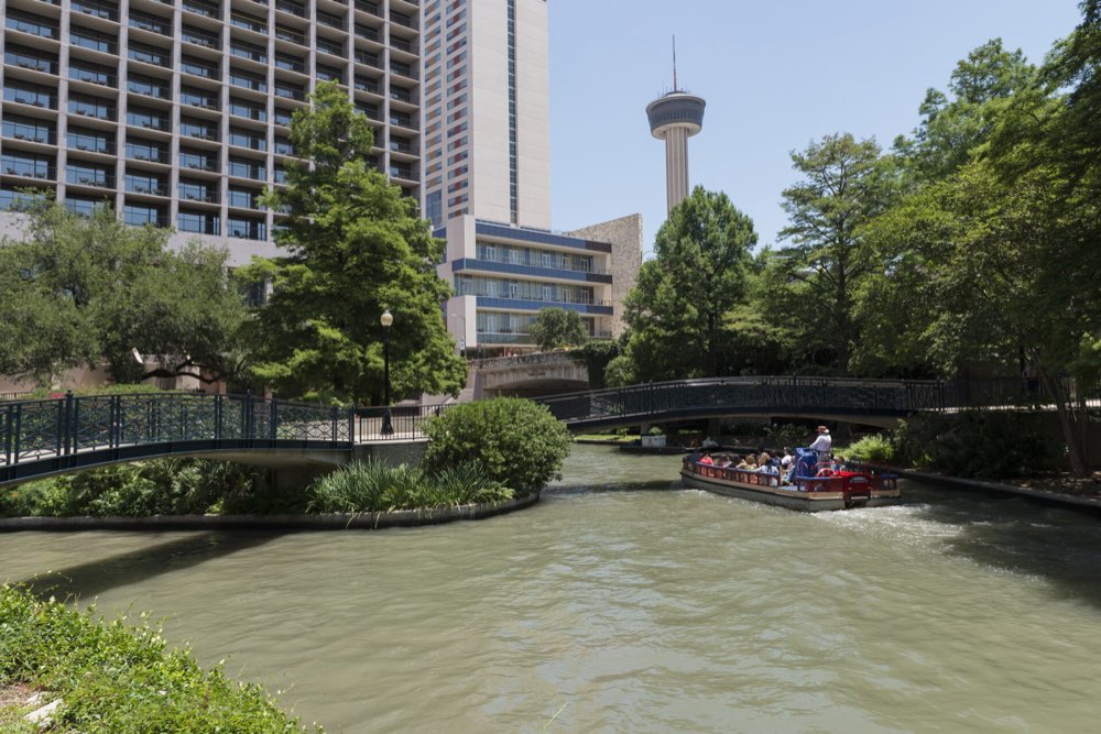 A tour barge with passengers rounds a bend of the San Antonio River Walk beneath footbridges, with Tower of the Americas rising above the trees