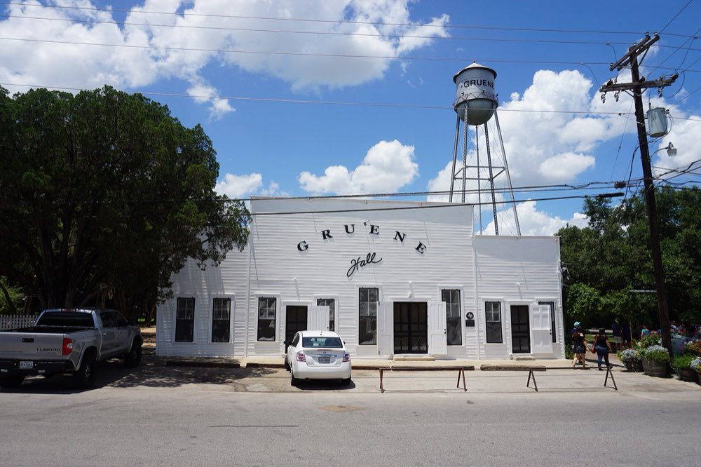 White clapboard Gruene Hall under a blue sky, with the Gruene water tower rising behind it