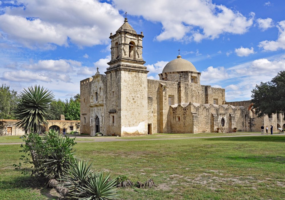 The limestone church of Mission San José with its bell tower and dome under a blue sky with white clouds, yucca plants in the foreground