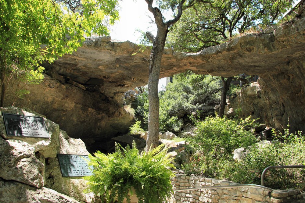 A limestone natural bridge arching over a shaded garden path, framed by oak trees and ferns