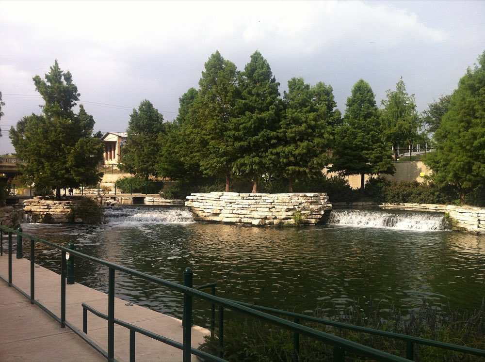 Small waterfalls and a calm pool at the north end of the Museum Reach beside Pearl, with railings and tall trees