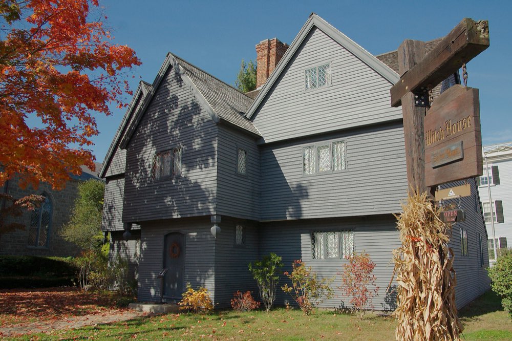 The steep-gabled, dark gray Witch House on a sunny autumn day, with a red-leaved tree on the left, a wooden hanging sign and a bundle of corn stalks on the right
