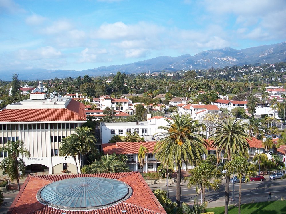 Red tile roofs, palm trees and white Spanish-style buildings of downtown Santa Barbara seen from above, with the Santa Ynez Mountains under a blue sky