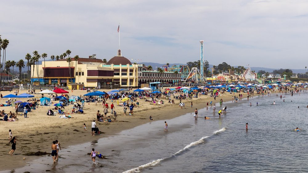 The Santa Cruz Beach Boardwalk seen across a wide sandy beach, with the domed Casino building, palm trees, rides and beach umbrellas along the shore