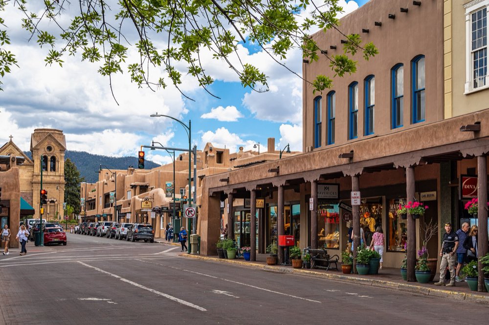 San Francisco Street in downtown Santa Fe, lined with adobe-style shops under wooden portals, with the cathedral's tower and green mountains at the far end under a blue sky