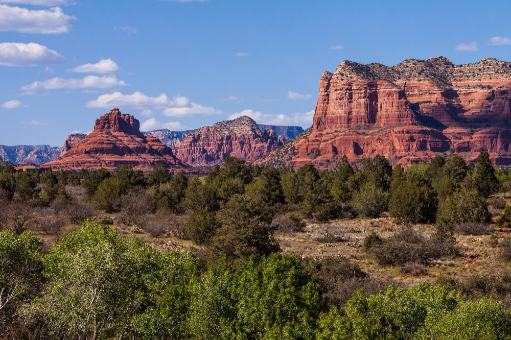 Bell Rock and the tall red cliffs of Courthouse Butte rising above green juniper and scrub under a blue sky with scattered clouds