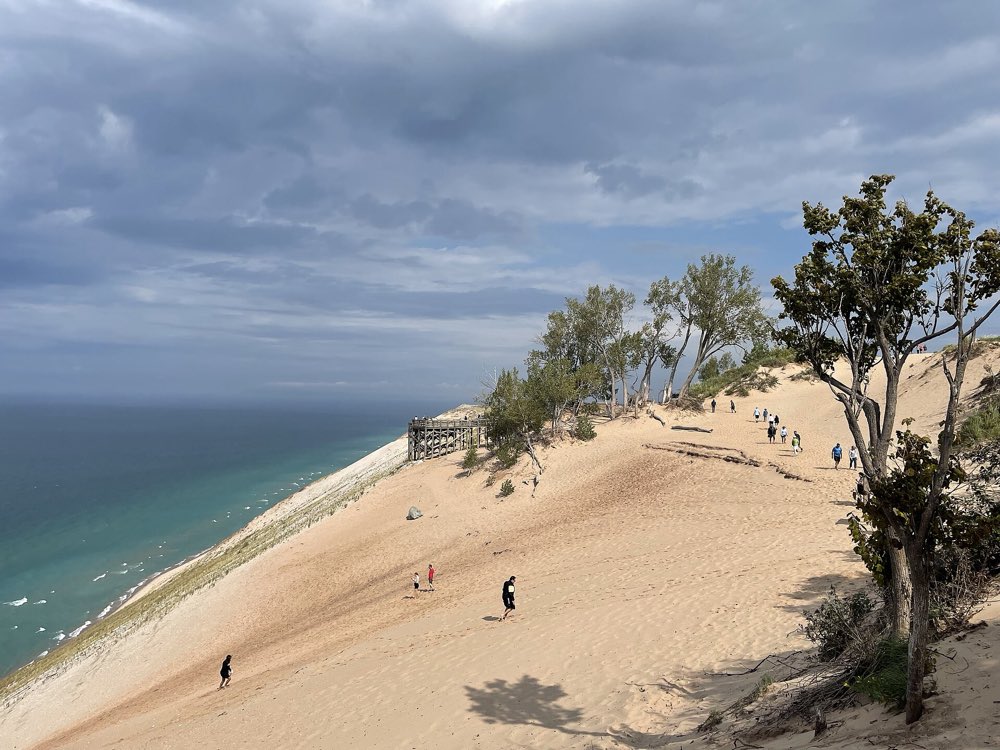 A huge sand bluff slopes down to turquoise Lake Michigan at Sleeping Bear Dunes, with a wooden overlook deck, a few small figures on the sand and windswept trees under a cloudy sky