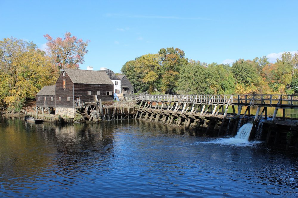 The weathered wooden gristmill and waterwheel at Philipsburg Manor beside a long wooden mill dam, with water spilling into the millpond and trees just turning gold and red behind