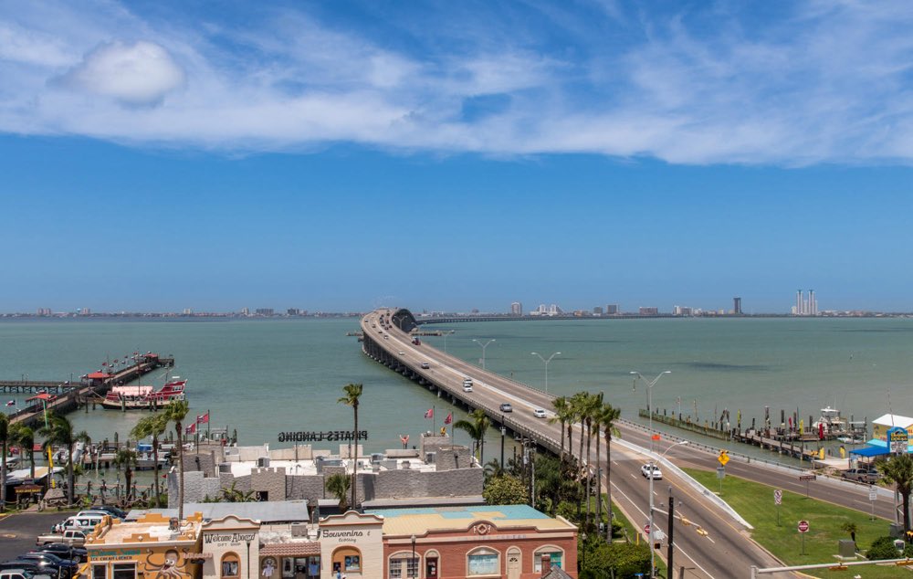 View from the top of the Port Isabel Lighthouse: old storefronts and palms in the foreground, the long Queen Isabella Causeway curving across green-blue water, and the South Padre Island skyline on the horizon