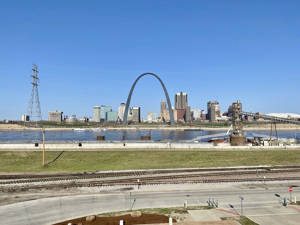 The steel Gateway Arch framing the downtown St. Louis skyline across the Mississippi River under a clear blue sky, seen from the Illinois bank