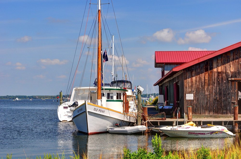 A white skipjack-rigged workboat with a tall wooden mast moored beside a weathered wooden boat shed with a red roof, on calm blue water at the Chesapeake Bay Maritime Museum