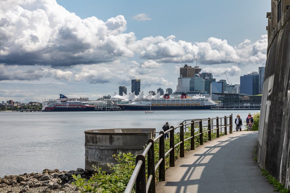 The paved Stanley Park seawall path curves along the water beside a stone wall, with cruise ships at Canada Place and the downtown Vancouver skyline across the harbour under big white clouds