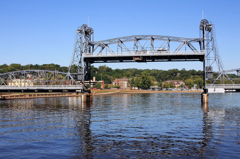The steel truss Stillwater Lift Bridge with its center span raised between two towers over the calm St. Croix River, wooded bluffs and town buildings behind under a clear blue sky