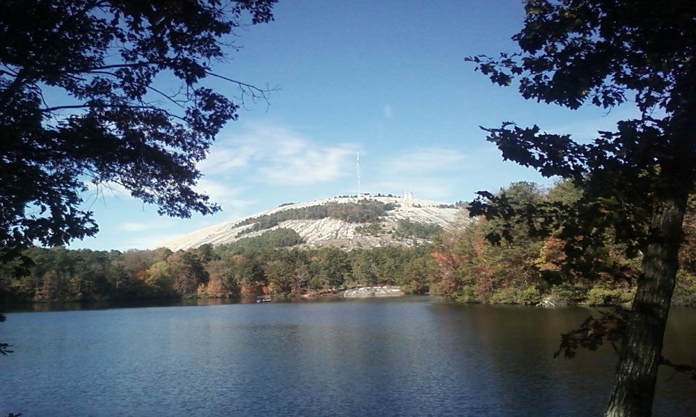 The bare granite dome of Stone Mountain rising above a lake ringed with autumn trees, under a blue sky with thin clouds