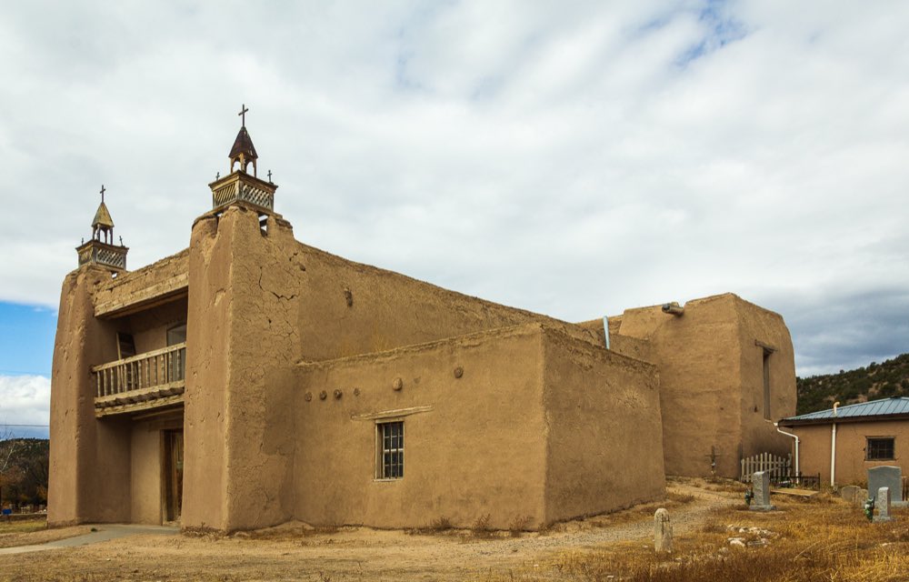 The twin wooden belfries and buttressed adobe walls of San José de Gracia church at Las Trampas, with weathered crosses in the dry grass of the churchyard under a pale sky