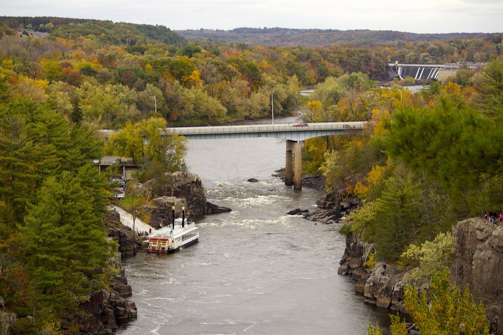 Looking down the rocky gorge of the St. Croix River from a cliff top, a white paddlewheel boat moored at a dock below, a highway bridge crossing the river and hills of orange and yellow autumn trees beyond