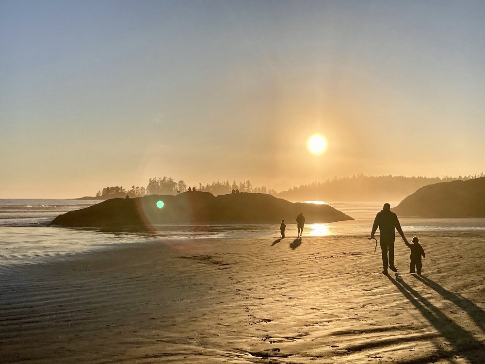 The low sun sets over the wet sand of Long Beach, with a rocky, tree-topped islet offshore and a few walkers in silhouette along the shore