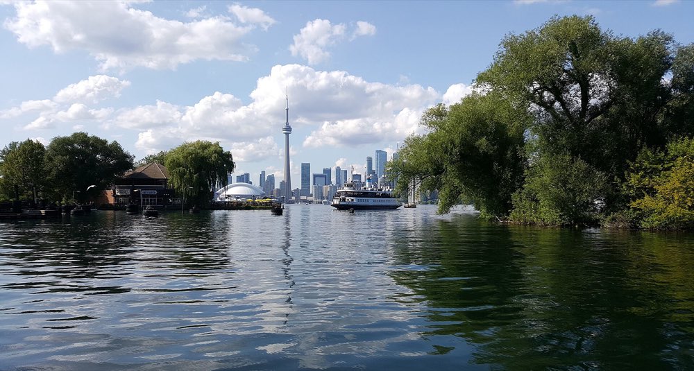 The Toronto skyline and CN Tower across calm lagoon water from Centre Island, with a white island ferry crossing between willow trees