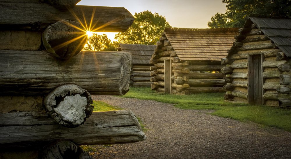 Low sun shining between reconstructed log huts along a gravel company street at Valley Forge