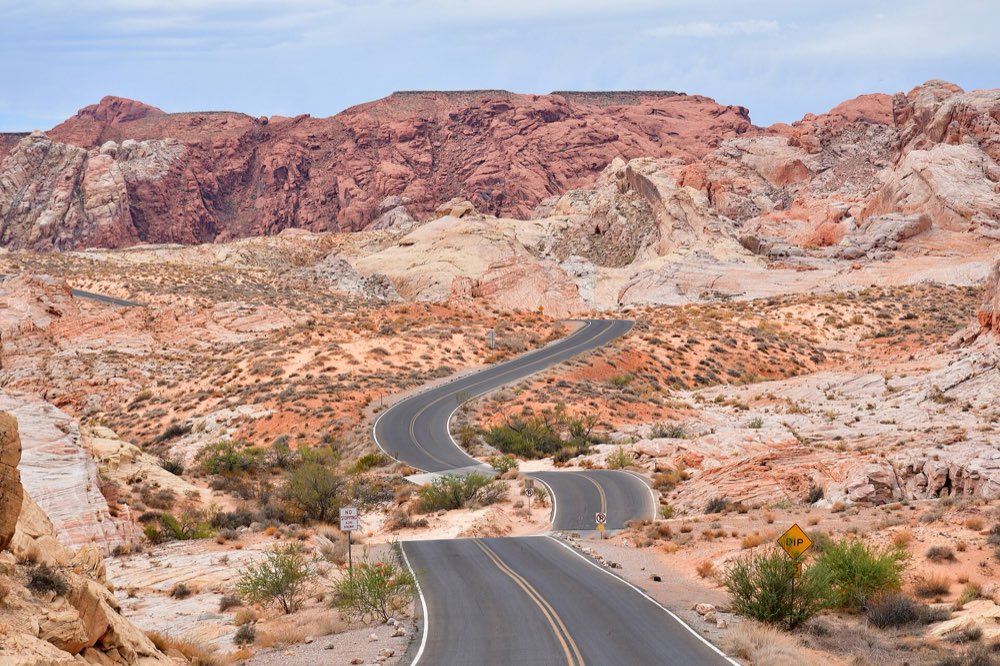A two-lane road winding through cream and orange sandstone toward a ridge of deep red rock under a pale cloudy sky