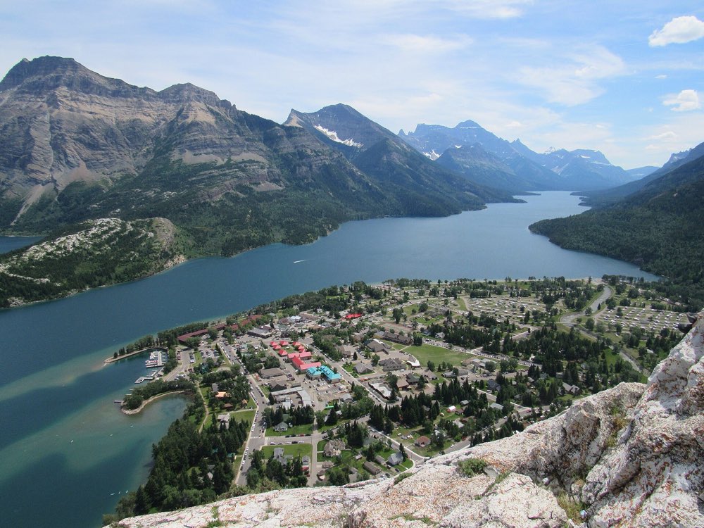 Looking down from a rocky summit on the Waterton townsite, with Upper Waterton Lake running south between steep mountains under a blue sky