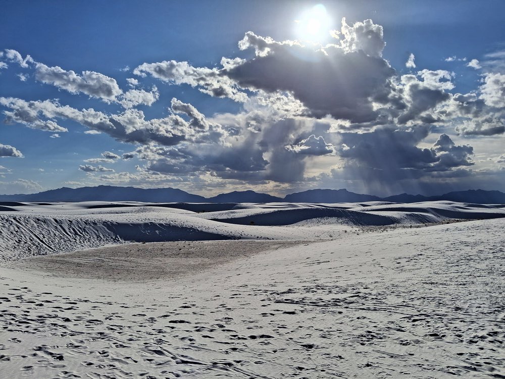 Rippled white gypsum dunes stretching to a line of dark mountains under a bright sun and heaped white clouds at White Sands National Park
