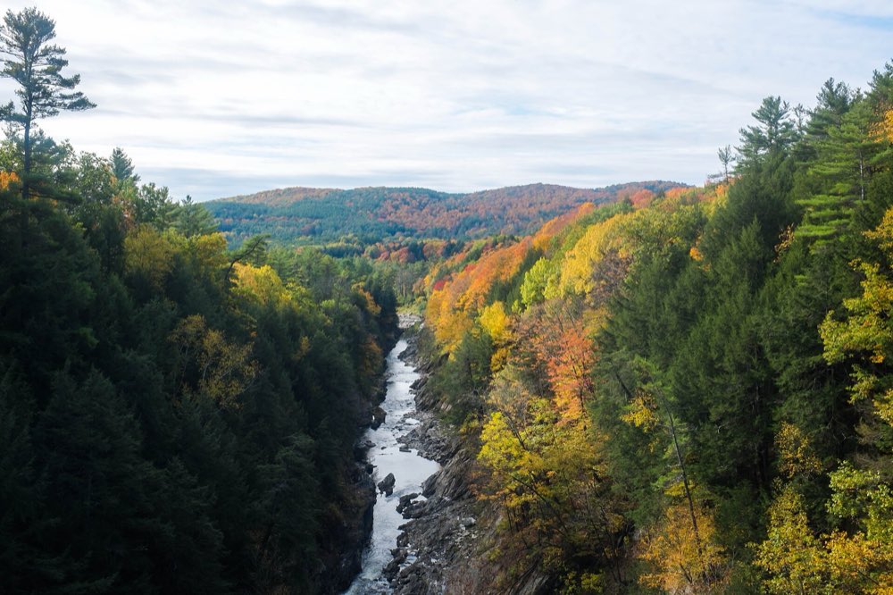 Looking down into Quechee Gorge from above, the Ottauquechee River running over rocks between steep slopes of dark pines and yellow, orange and red maples, with a hazy ridge beyond