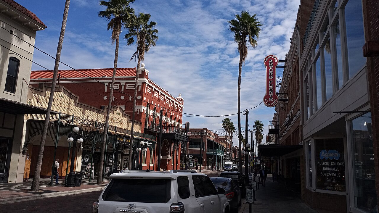 Palm-lined 7th Avenue in Ybor City, with red-brick historic buildings, iron balconies and a vertical cigar sign under a blue sky